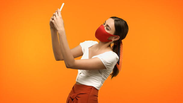 Woman in protective mask taking photo on smartphone isolated on orange  - Фото, изображение