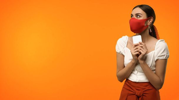 Koruyucu maskeli genç bir kadın portakalda izole bir cep telefonu tutuyor.  - Fotoğraf, Görsel