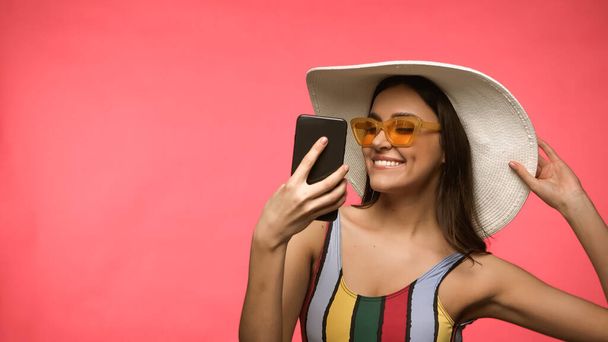 Cheerful woman in sun hat and swimwear holding mobile phone isolated on pink  - Foto, afbeelding