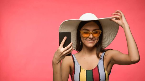Happy woman in sun hat and swimwear holding cellphone and looking at camera isolated on pink  - Fotó, kép