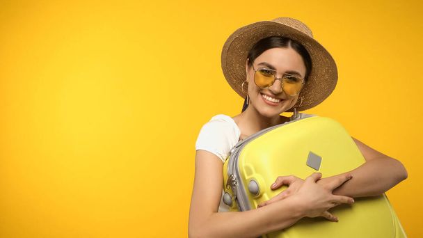 Glückliche Frau mit Strohhut umarmt Koffer isoliert auf gelb  - Foto, Bild