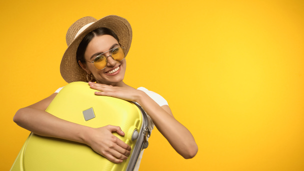 Jeune femme en lunettes de soleil étreinte valise isolée sur jaune  - Photo, image