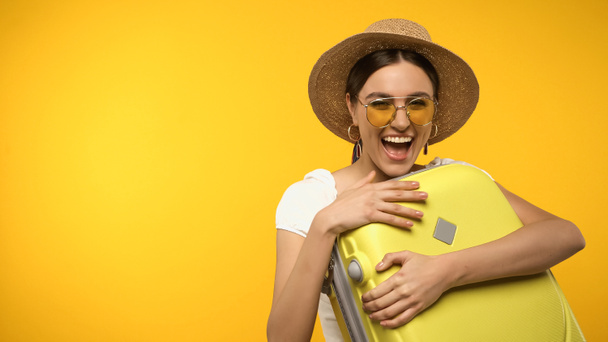 Excited woman in sun hat hugging suitcase isolated on yellow  - Zdjęcie, obraz