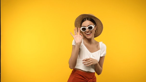 Smiling μοντέλο σε γυαλιά ηλίου και ψάθινο καπέλο κουνώντας το χέρι απομονώνονται σε κίτρινο  - Φωτογραφία, εικόνα