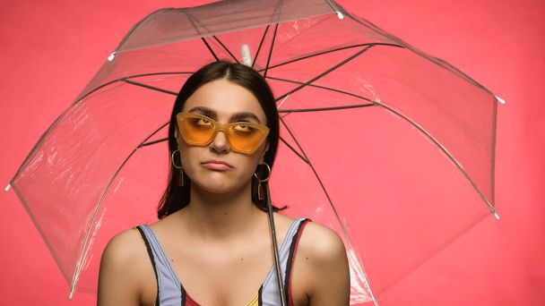 Disappointed woman in swimsuit and sunglasses looking at umbrella isolated on pink  - Φωτογραφία, εικόνα