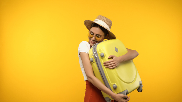 Happy tourist in straw hat closing eyes while hugging suitcase isolated on yellow  - Photo, Image