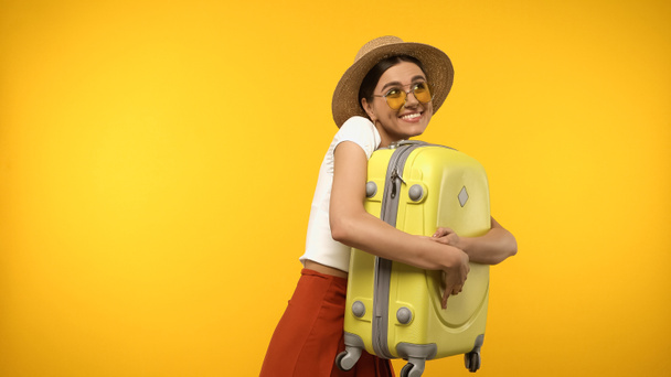 Traveler in sunglasses and straw hat hugging suitcase isolated on yellow  - Фото, зображення