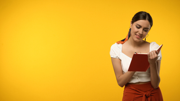 Cheerful model in blouse writing on notebook isolated on yellow - Фото, изображение