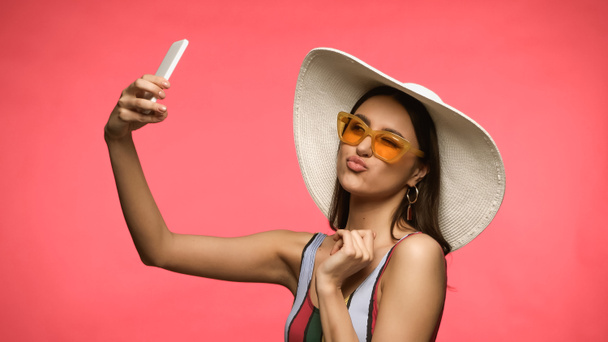 Brunette woman in sun hat and sunglasses taking selfie on smartphone isolated on pink  - Fotó, kép