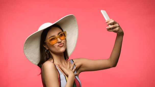 Young woman in straw hat and swimsuit taking selfie on smartphone and grimacing isolated on pink  - Photo, image