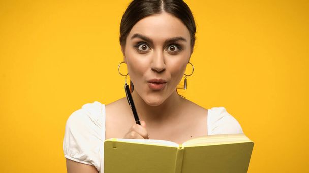 Excited woman holding pen near notebook isolated on yellow - Photo, Image