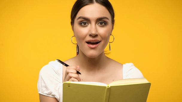Amazed brunette woman holding pen near notebook isolated on yellow - Фото, изображение