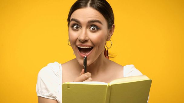 Excited woman in blouse holding pen near notebook isolated on yellow - Foto, Imagem