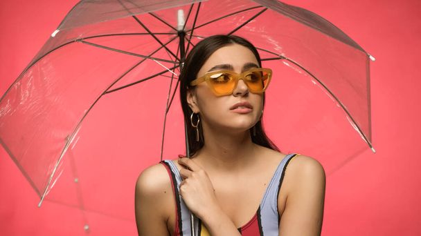 Young woman in sunglasses and swimsuit holding umbrella isolated on pink  - Foto, Imagen