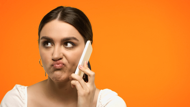 verwarde brunette vrouw grimacing tijdens het praten op smartphone geïsoleerd op oranje  - Foto, afbeelding