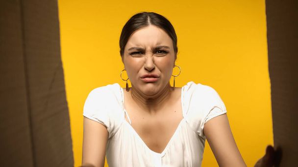 Bottom view of disgusted brunette woman looking at open cardboard box isolated on yellow  - Photo, Image