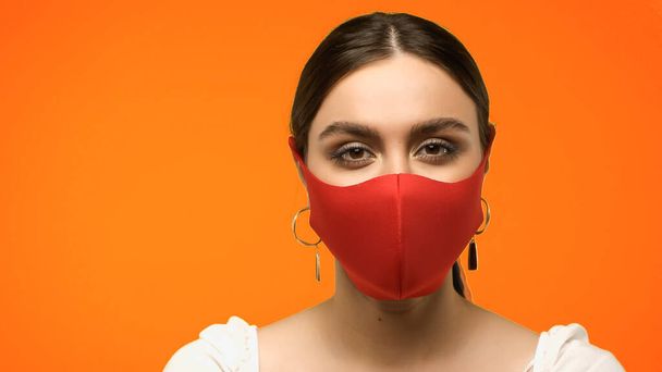 Молода жінка в захисній масці і сережки дивиться на камеру ізольовано на помаранчевому
  - Фото, зображення