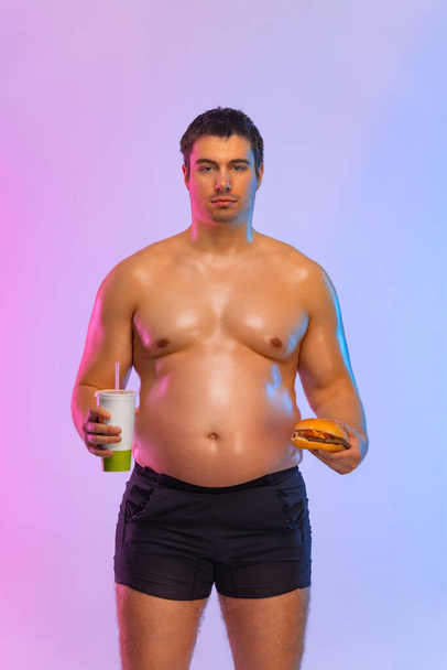 Fat man with burger and soda. Not sporty men drinking and eating hamburger isolated on pink background. - Fotó, kép