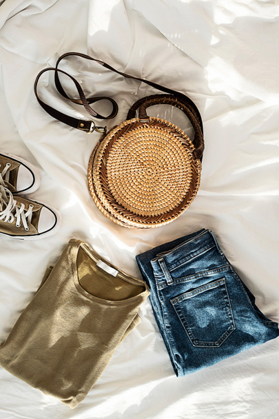 Blue jeans, trendy round rattan woven bag, khaki shirt and sneakers on white bedding. Women's fashion spring or summer outfit. Stylish clothes collage, top view. - Foto, Imagen