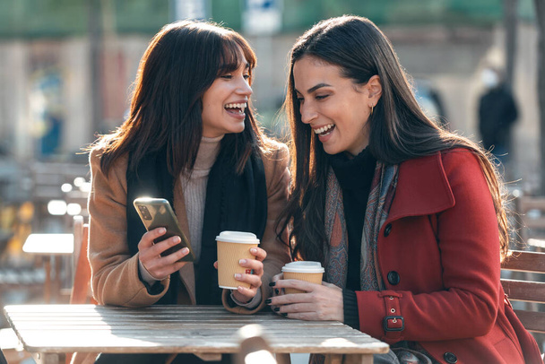 Shot of two attractive friends enjoying coffee together while looking smartphone and talking sitting on the terrace of a bar. - 写真・画像