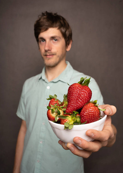 Young handsome tall slim white man with brown hair offering you bunch of strawberries in light blue shirt on grey background - Fotó, kép