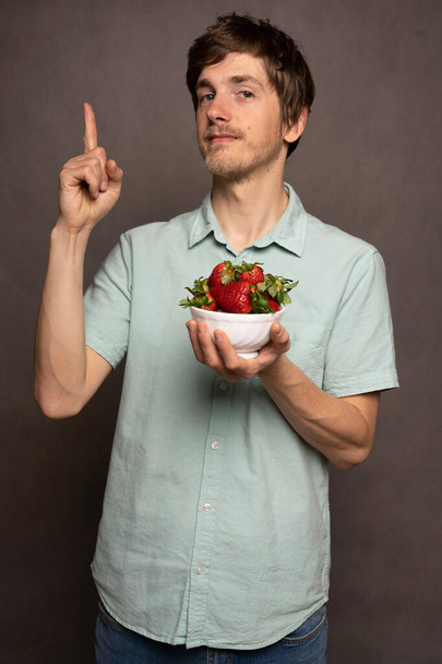 Young handsome tall slim white man with brown hair holding strawberries pointing up in light blue shirt on grey background - Fotografie, Obrázek