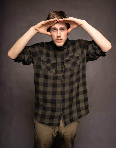 Young handsome tall slim white man with brown hair trying to see with flannel shirt on grey background - Фото, изображение