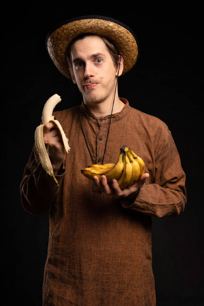 Young handsome tall slim white man with brown hair eating banana licking his lips with brown shirt and straw hat on black background - Fotó, kép