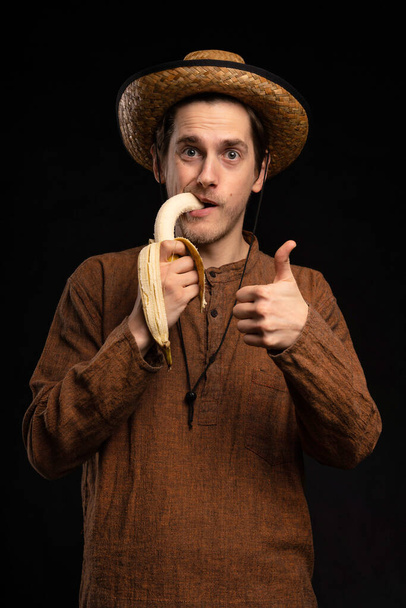 Young handsome tall slim white man with brown hair biting banana thumbs up with brown shirt and straw hat on black background - Fotografie, Obrázek
