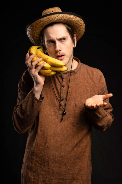 Young handsome tall slim white man with brown hair using bananas as phone looking concerned with brown shirt and straw hat on black background - Photo, Image
