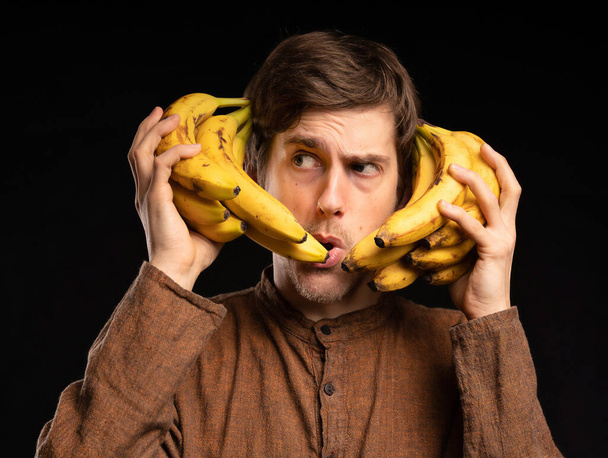 Young handsome tall slim white man with brown hair holding bananas looking curious with brown shirt on black background - Foto, Imagen