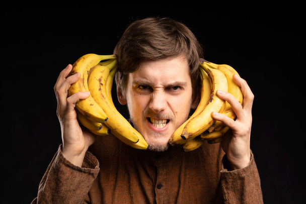 Young handsome tall slim white man with brown hair holding bananas next to his face looking angry with brown shirt on black background - Fotó, kép