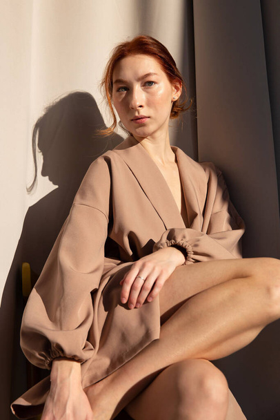 Young european woman with white skin with curtains behind in beige clothes - Foto, afbeelding