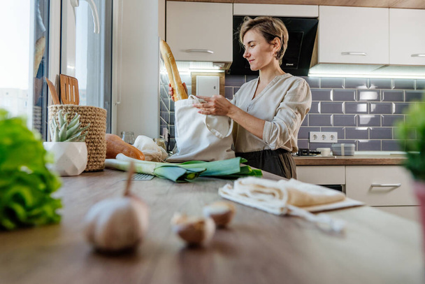 Mujer joven sosteniendo bolsa reutilizable de algodón con comida en la cocina. Cero residuos y concepto de embalaje sostenible. - Foto, imagen