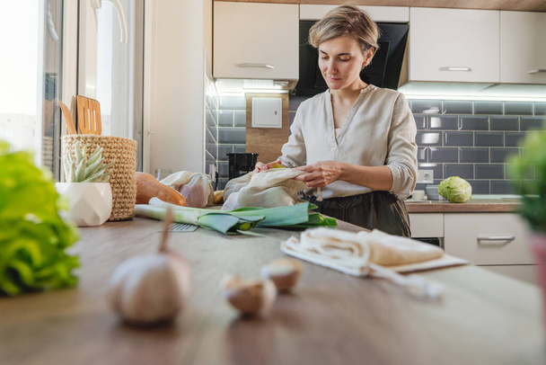 Young woman pulls groceries out of the mesh bag in the kitchen. Food in the organic cotton reusable produce bag. Zero waste and sustainable packaging concept. - 写真・画像