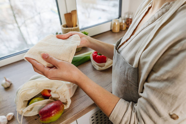 Young woman tie the organic cotton reusable produce bag with vegetables and fruits on the table in the kitchen. Zero waste and sustainable packaging concept. - Foto, Imagem