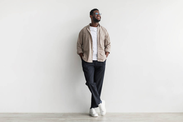 Pleine longueur de beau jeune homme afro-américain portant des vêtements décontractés, regardant de côté, posant contre un mur blanc - Photo, image