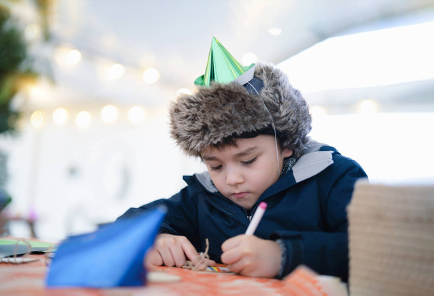 Cute boy with party hat using pink pen drawing or writing on paper colour with blurry bright light background, Kid having fun at birthday party, Child doing activity on Christmas or New Year holiday - Foto, Imagem