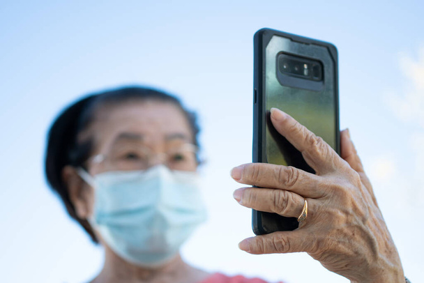 Glückliche asiatische Senioren mit Gesichtsmaske, die mit ihrem Smartphone im Park einen Videoanruf entgegennehmen und mit Familie, Technik und älteren Menschen chatten.. - Foto, Bild