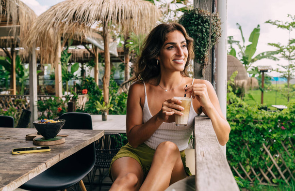 Positive ethnic female in casual clothes sitting at table in cafe and drinking glass of cocktail while looking away in summer day - Foto, imagen
