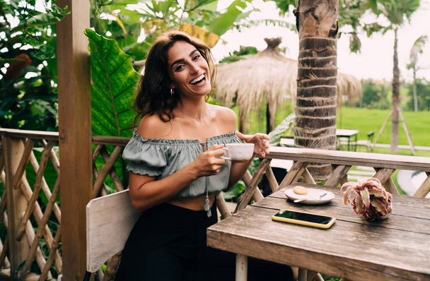 Delighted young ethnic woman in stylish summer clothes sitting on veranda of cafe in tropical garden and drinking coffee during breakfast - Fotó, kép