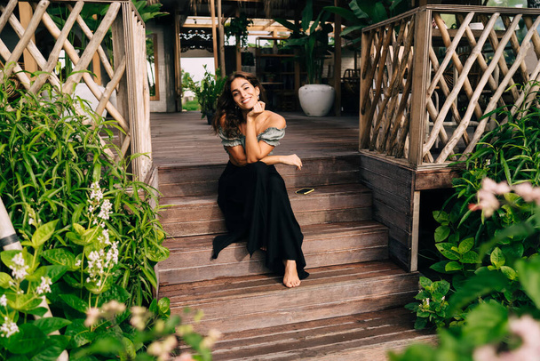 Full body smiling barefoot woman in trendy clothes sitting on cafe steps leaning chin on hand while admiring plants surrounding summer wooden terrace in sunny day - Photo, image