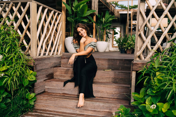 Full body smiling woman in trendy outfit sitting on steps of wooden terrace bending head to shoulder and leaning hands on knee while enjoying plants decorating entrance to summer cafe against plants decking interior of room in sunny day - Photo, Image