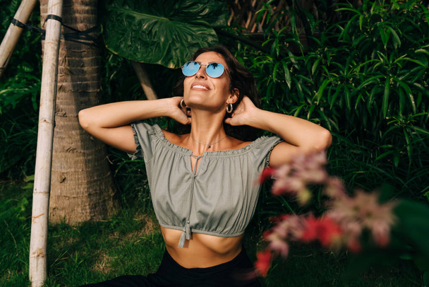Positive ethnic young slim female traveler in summer clothes and sunglasses touching hair and looking up while recreating on green grass in exotic garden - Φωτογραφία, εικόνα
