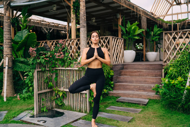 Calm female in sportive tracksuit standing in tree pose and pray during morning meditation at villa patio, Caucasian woman enjoying yoga practice for mindfulness and enlightenment harmony - Photo, image