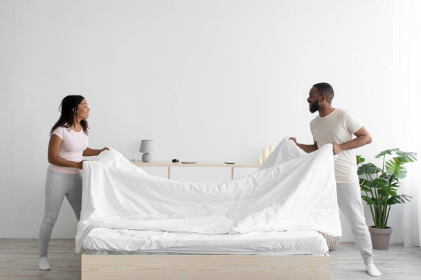 Vrolijke jonge zwarte vrouw en man maken bed en houden deken in slaapkamer interieur - Foto, afbeelding