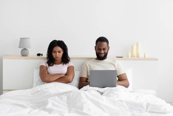 Offended sad young black woman angry at husband with computer, sit on bed in bedroom interior - Photo, image