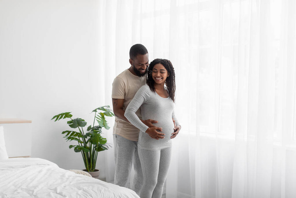 contento sonriendo millennial africano americano chico en doméstica ropa abraza su embarazada mujer, toca vientre - Foto, Imagen