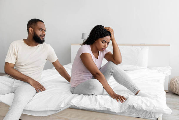Serious young black man calm to sad desperate depressed wife, sit on bed in bedroom interior - Фото, изображение