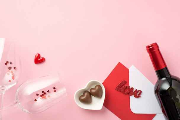 Top view photo of valentine's day decor wineglasses with confetti heart shaped saucer with candies envelope red card inscription love and wine bottle on isolated pastel pink background with copyspace - 写真・画像